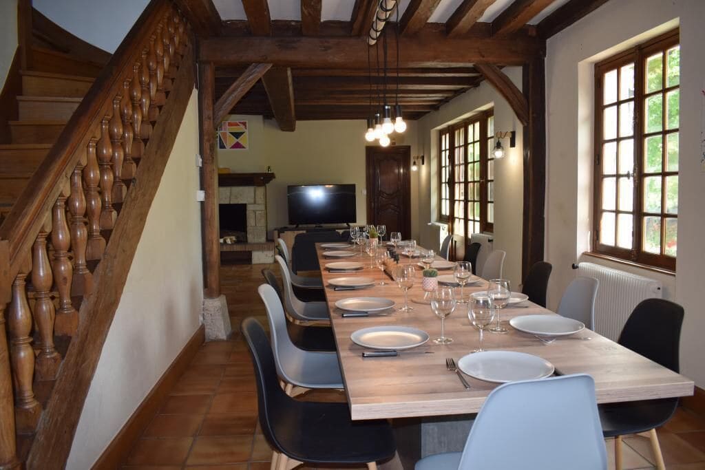 A dining area with a long wooden table set for six, surrounded by chairs, a staircase, and a television in the background.