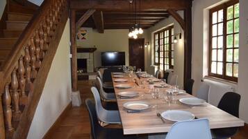 A dining area with a long wooden table set for six, surrounded by chairs, a staircase, and a television in the background.