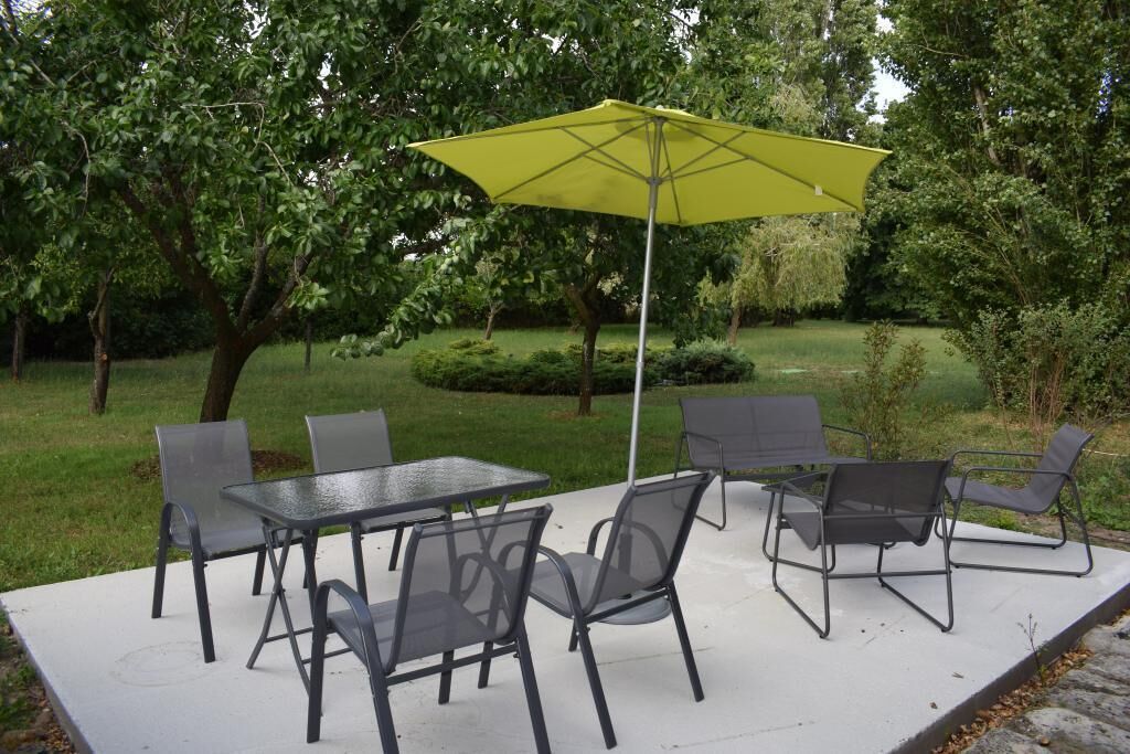 Un patio al aire libre con una mesa, sillas, un paraguas amarillo y abundante vegetación.
