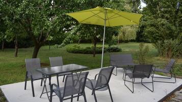 Un patio al aire libre con una mesa, sillas, un paraguas amarillo y abundante vegetación.