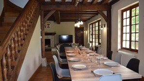 Comedor con una larga mesa de madera dispuesta para seis, rodeada de sillas, una escalera y un televisor al fondo.