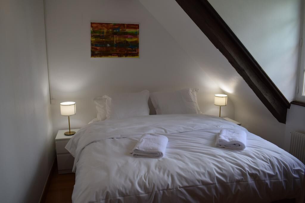 Een net opgemaakt bed met witte lakens en twee gevouwen handdoeken, een schilderij aan de muur en een schuine plafond.