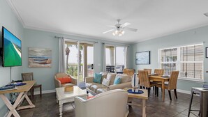 A spacious living room with a large sofa, dining table, and chairs. There are large windows with blinds, a ceiling fan, and artwork on the walls.
