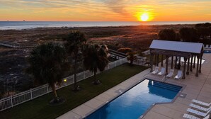 A pool area with a view of the sunset over the ocean, palm trees, and a covered pavilion with lounge chairs.