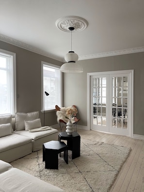 Un salon comprenant un canapé, une table basse, un vase avec des fleurs, un luminaire suspendu et une vue sur un espace extérieur, accessible par une porte vitrée.