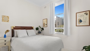 Quarto com uma cama grande, mesa de cabeceira, janela com cortinas e uma obra de arte na parede.