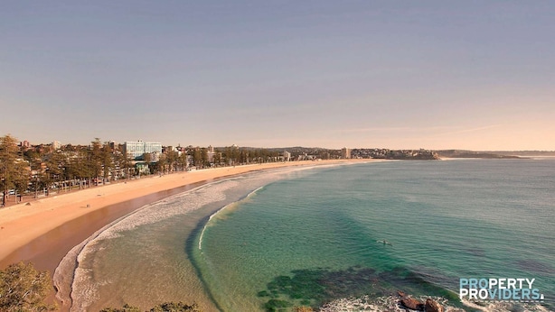 Uma praia costeira com águas cristalinas, areal extenso e uma fileira de edifícios ao longo da costa.