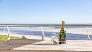 A bottle of Cuvée Brut champagne and a glass on a wooden deck with a beach and ocean in the background.