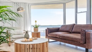A modern living room with a leather sofa, a round wooden coffee table with snacks, a potted plant, and a view of the ocean through large windows.