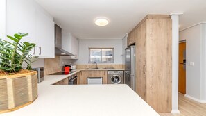 A modern kitchen with white cabinets, a wooden accent wall, a washing machine, and a built-in oven. - Lagoon Beach Gem Pool, Views & sea breeze (Milnerton)