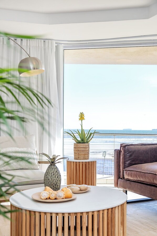A modern living room with a round wooden coffee table, a brown leather sofa, a potted plant, and a view of the ocean.