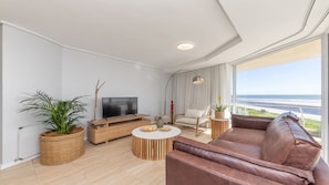 A modern living room with a leather sofa, a wooden TV stand, a round coffee table, and a large window with a view of the beach.