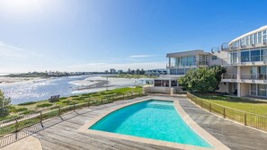 Ein Pool mit Holzterrasse, Blick auf den Strand und moderne Gebäude im Hintergrund.