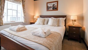 A neatly made bed with white linens and a floral-patterned duvet, two bedside lamps, a framed picture on the wall, and a window with curtains. - Midships Cottage (Whitby)