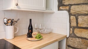 A kitchen with white cabinets, a stone wall, a white mug with utensils, a bottle of wine, and a plate with grapes and cheese. - Midships Cottage (Whitby)
