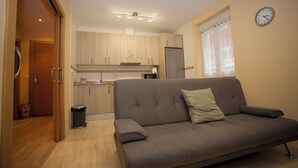 A modern living room with a grey sofa, a small trash bin, and a kitchen area with wooden cabinets and a refrigerator.