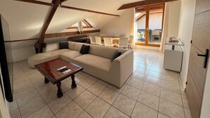 A spacious living area with a large L-shaped sofa, a wooden coffee table, and a dining table with chairs. There is a kitchen area with a refrigerator and a dining table visible through a glass door. - La Parenthèse Enchantée (Sevrier)