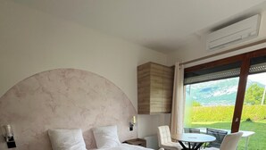A bedroom with a large bed, a wooden headboard, a nightstand, a chair, a table, and a view of a garden and mountains through a window. - La Parenthèse Enchantée (Sevrier)