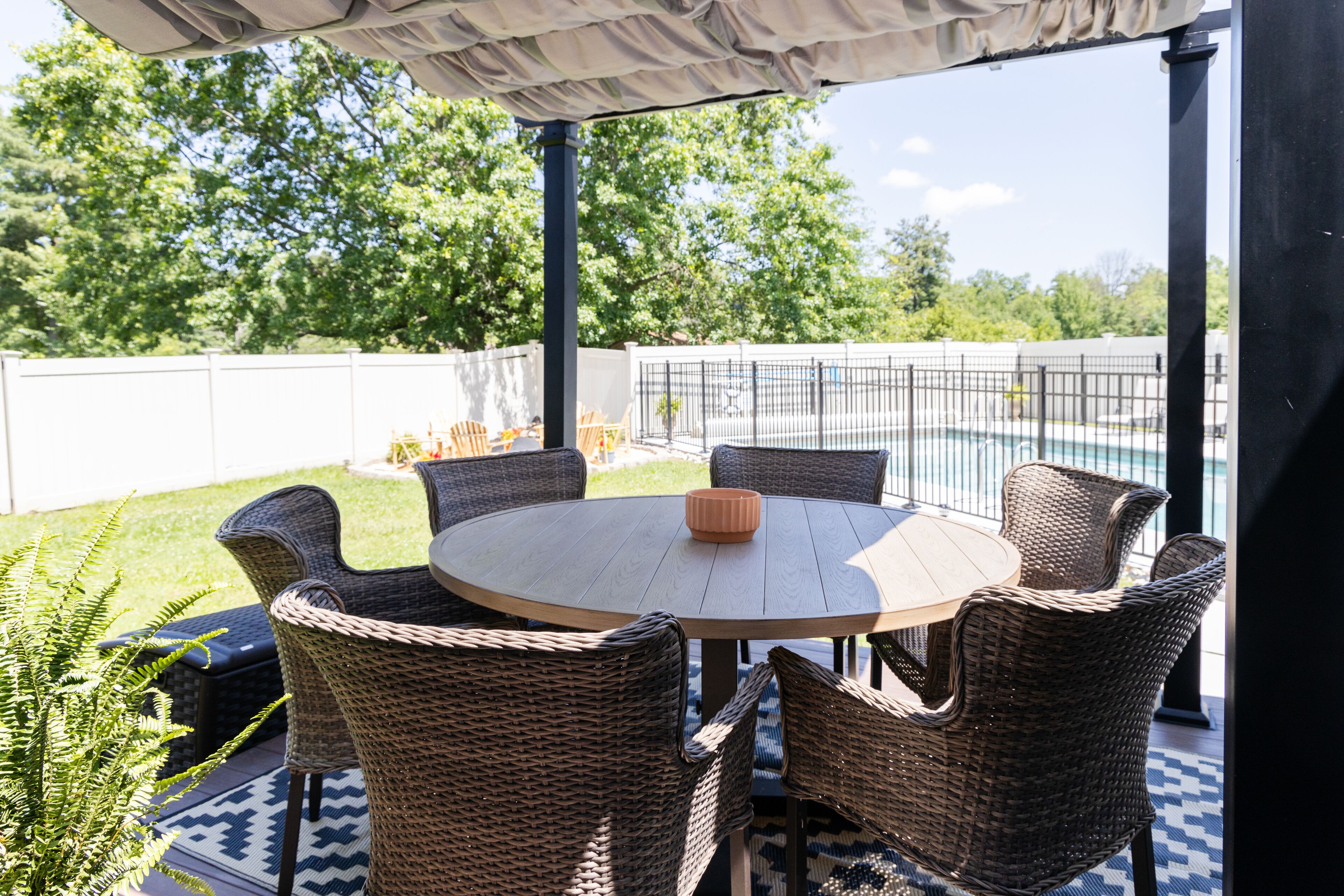 A covered patio with a round table and wicker chairs, a pool in the background, and a white fence.