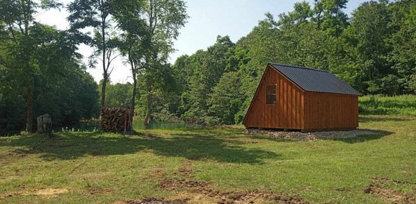 Una cabaña de madera pequeña con techo inclinado, rodeada por un campo cubierto de hierba y árboles frondosos.
