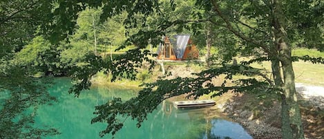 A cabin with a sloped roof, surrounded by dense trees and a calm lake.