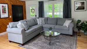 A living room with a grey sectional sofa, a glass coffee table, a patterned rug, a door, a window with curtains, and a framed picture on the wall.