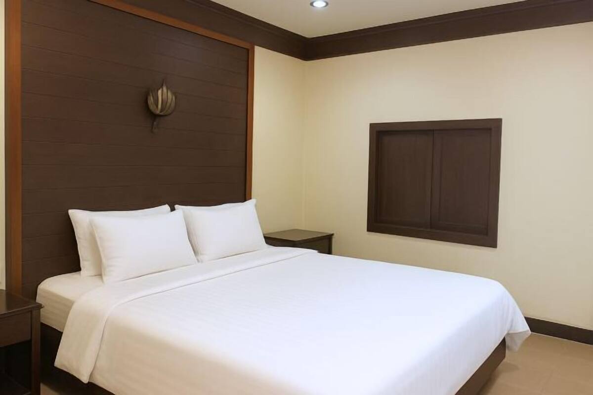 A neatly made bed in a hotel room with a wooden headboard, a small bedside table, and a window with a dark frame.