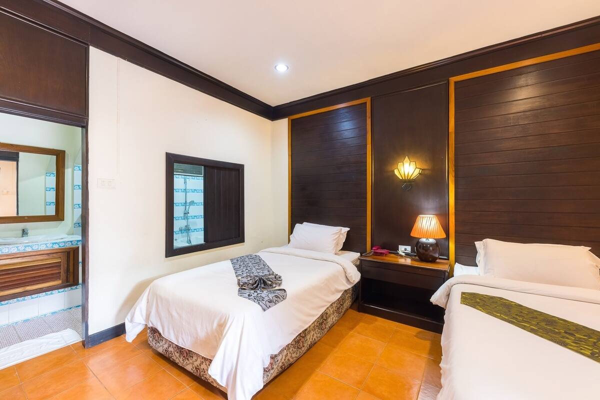 A hotel room with two beds, a large mirror, a window, a nightstand with a lamp, and a bathroom with a sink and mirror.