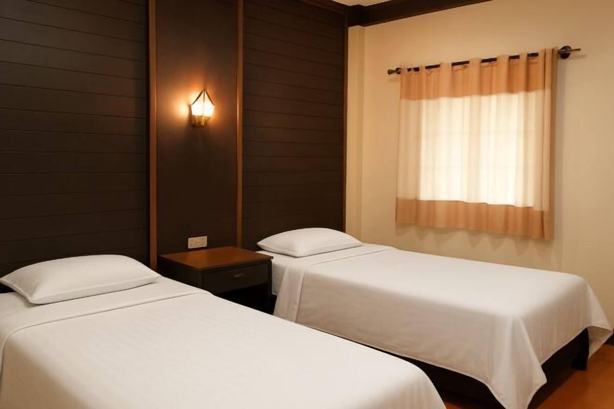 A room with two single beds, a nightstand, a wall-mounted light, and a window with curtains.