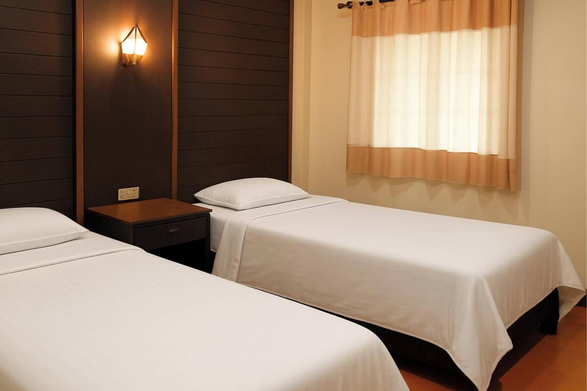 A hotel room with two single beds, a nightstand, a wall-mounted light, and a window with curtains.