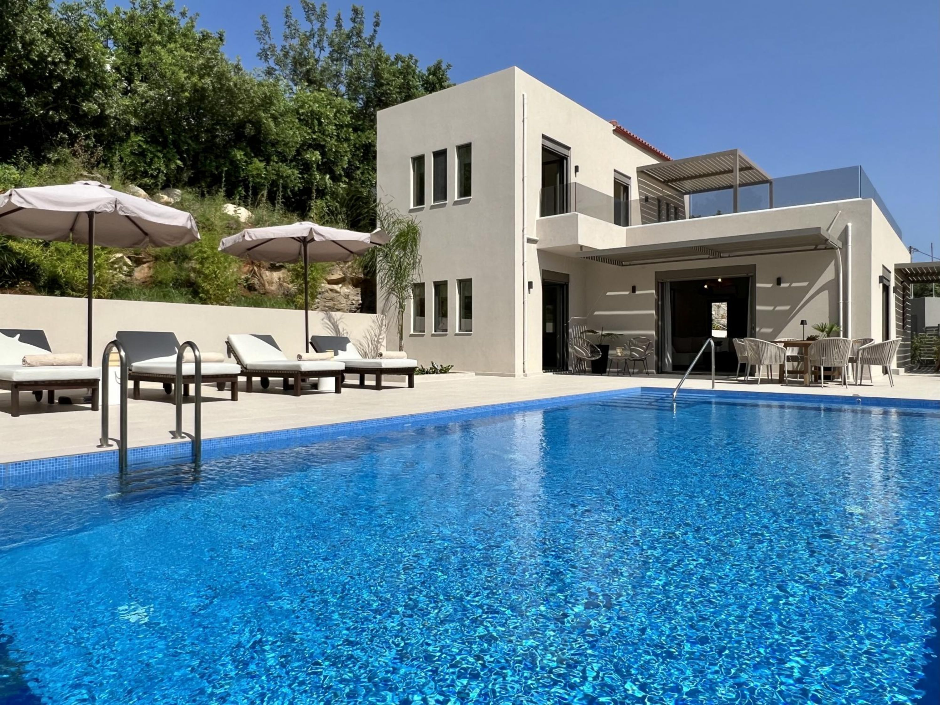 Un espace moderne aménagé au bord de la piscine, avec des chaises longues et des parasols, une piscine à l’eau d’un bleu éclatant et un bâtiment à deux étages doté de grandes fenêtres.