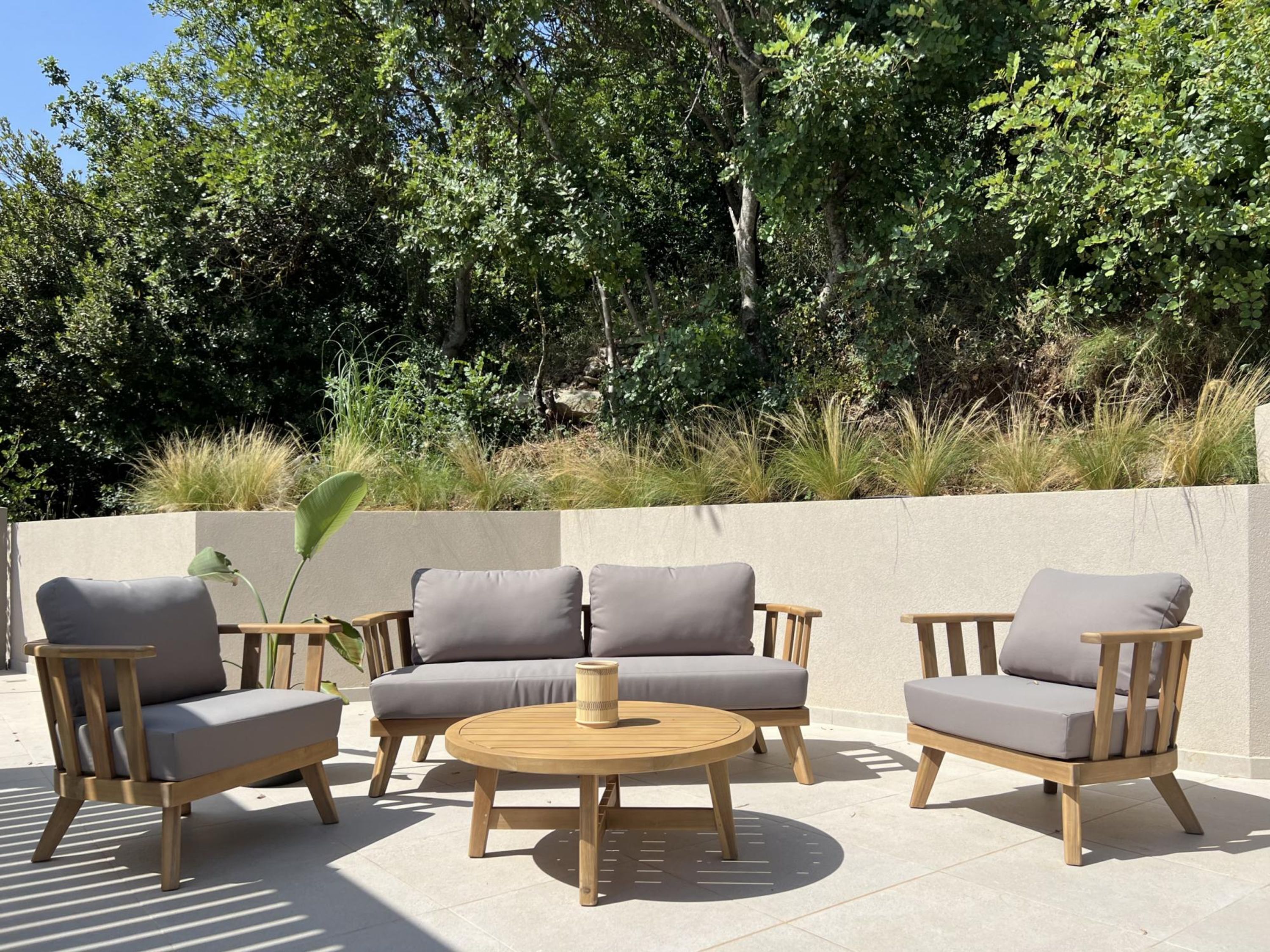 Un espace extérieur aménagé avec une table ronde en bois et trois chaises rembourrées, entouré d’un petit muret et d’une végétation luxuriante.