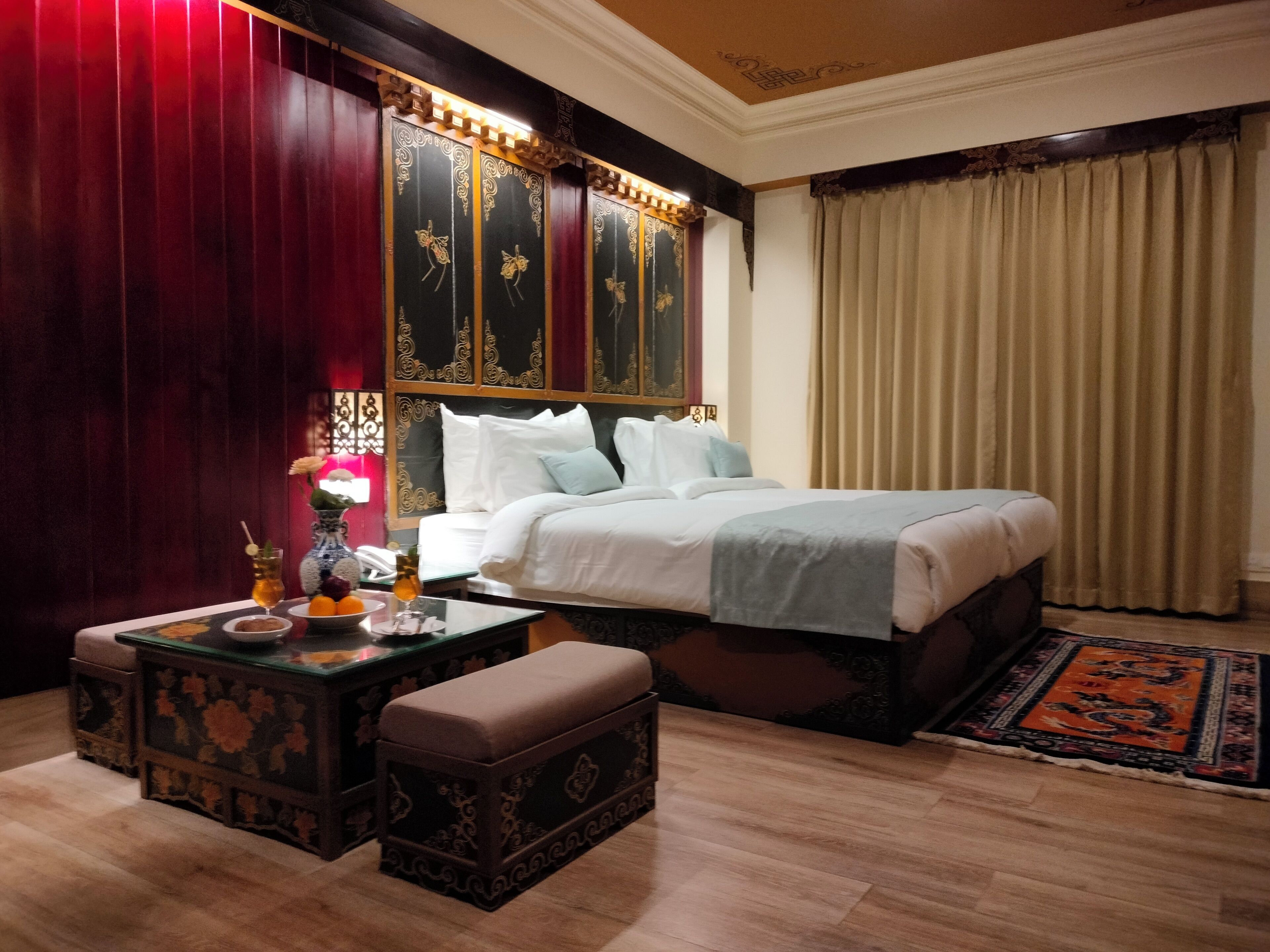 Habitación de hotel con una cama grande, una mesita de noche con una lámpara, una pequeña mesa con un florero y un teléfono, además de un banco decorativo.