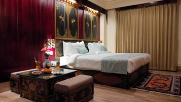 Habitación de hotel con una cama grande, una mesita de noche con una lámpara, una pequeña mesa con un florero y un teléfono, además de un banco decorativo.