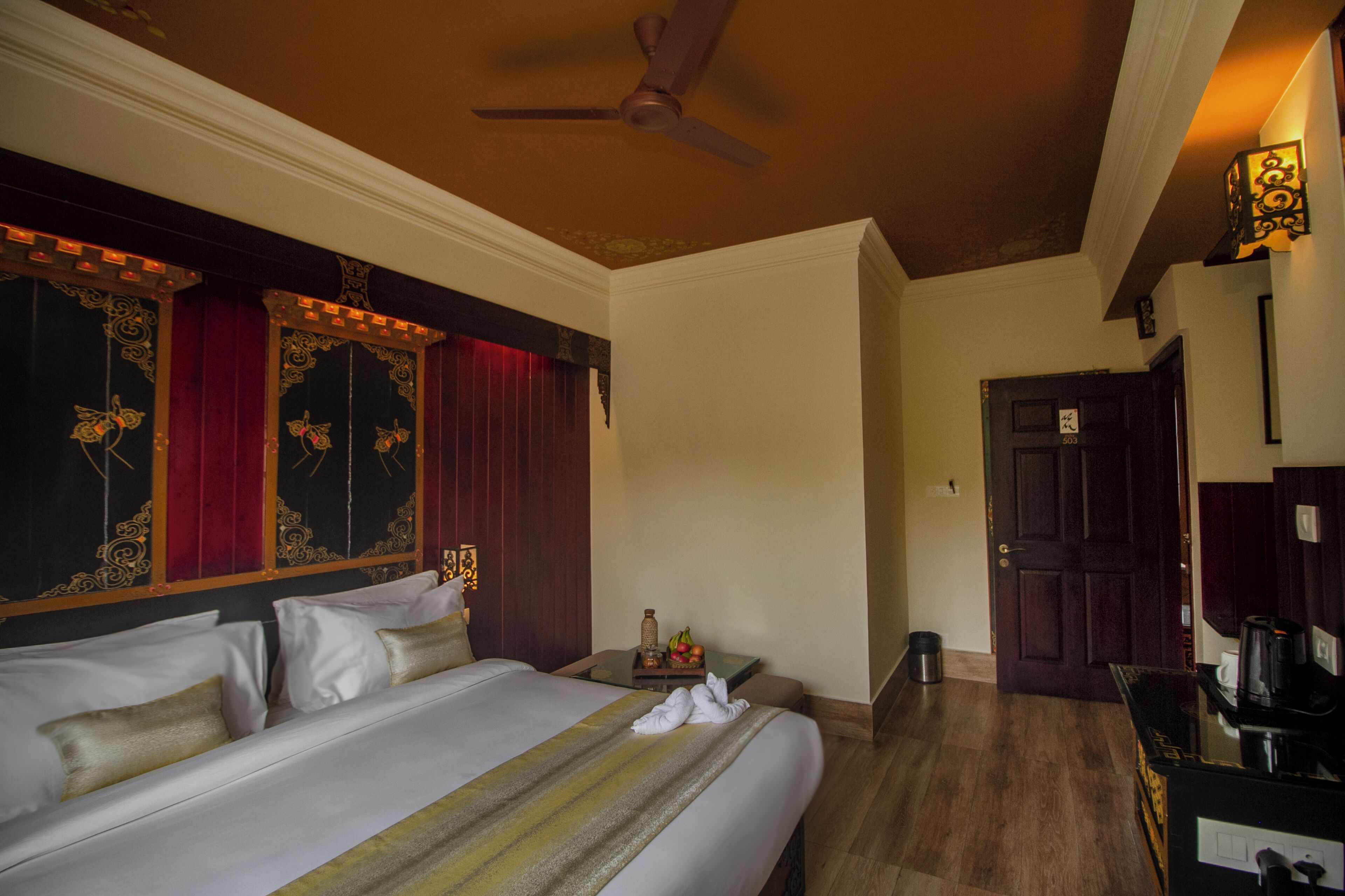 A hotel room with a large bed, a ceiling fan, a wooden door, and a small table with a bottle and fruit.