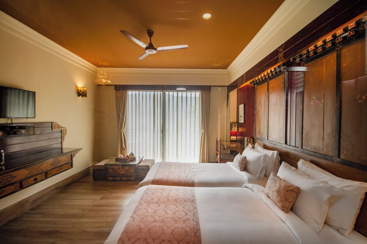 A hotel room with two beds, a ceiling fan, a television, and a large window with curtains.
