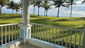 A balcony with a white railing overlooking a well-maintained lawn with palm trees and a view of the ocean.