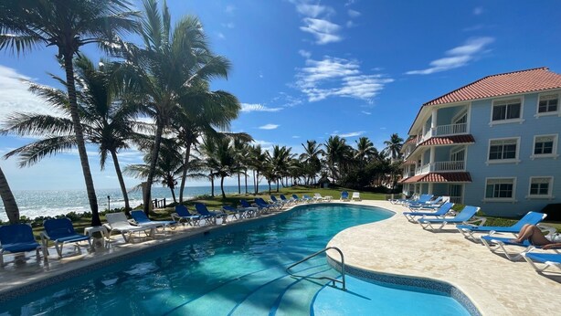 A pool with blue lounge chairs, palm trees, and a building with a red roof.