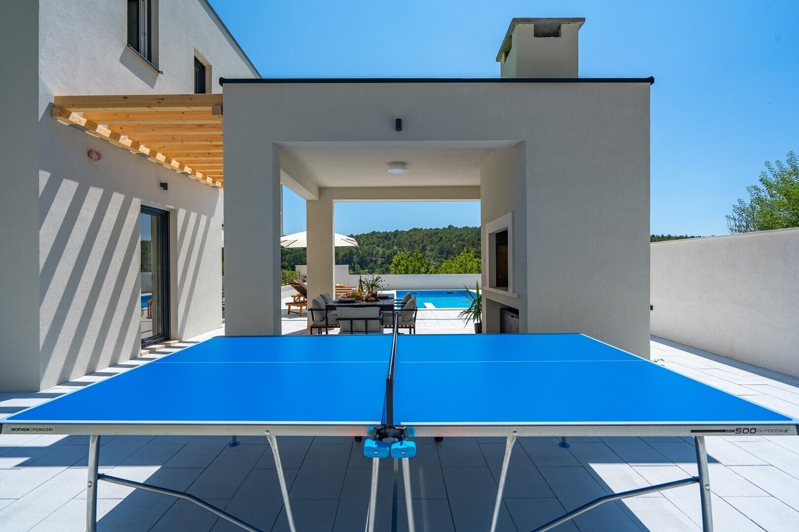 Een tafeltennistafel op een terras, met een zwembad en een zitgedeelte buiten op de achtergrond.