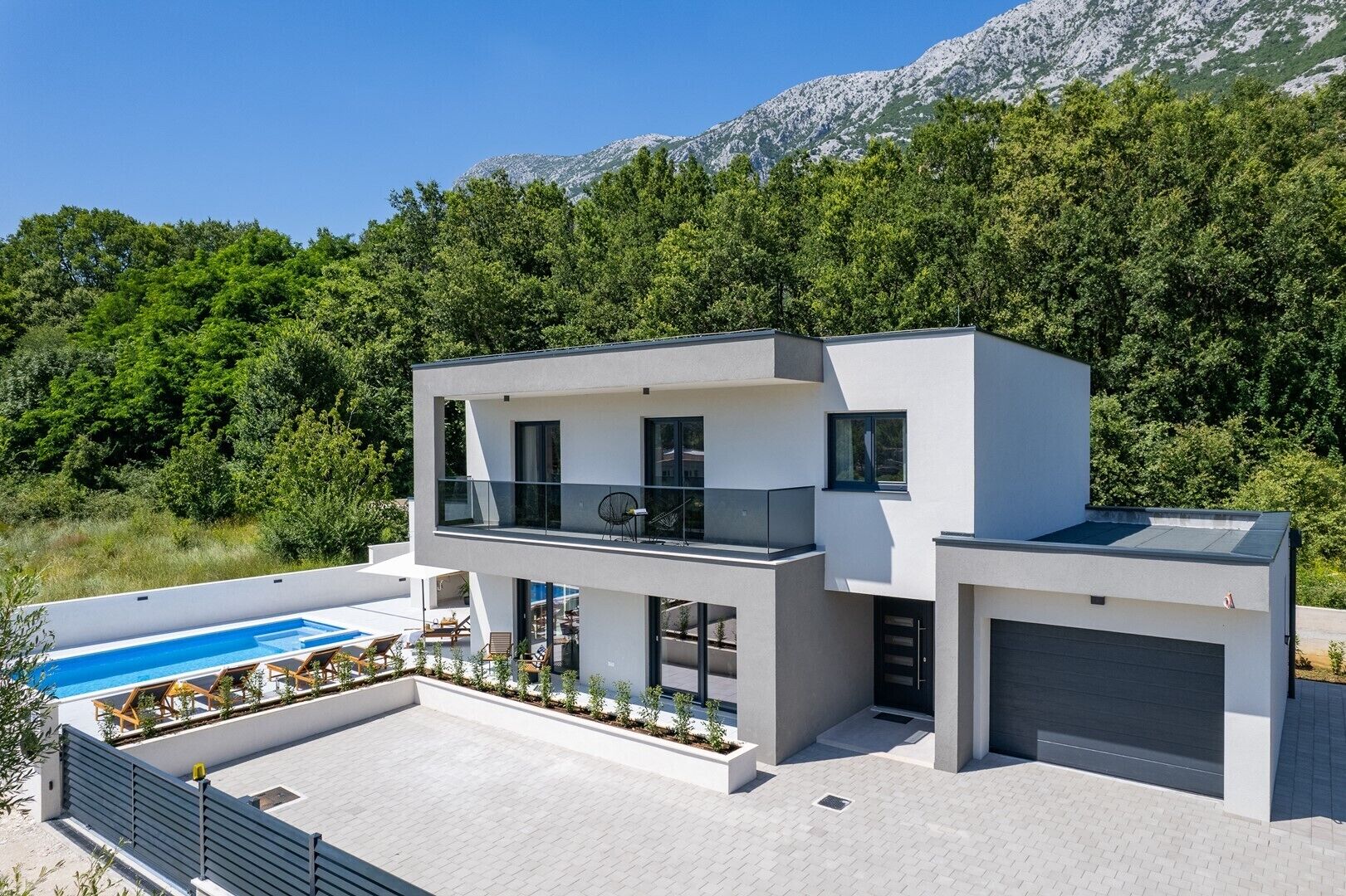 Una casa moderna de dos pisos con piscina, garaje y balcón con vista a un área boscosa y montañas al fondo.