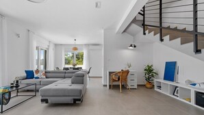 A modern living room with a grey sectional sofa, a dining area with a table and chairs, a white desk with a wicker chair, and a staircase with a metal railing. - Stylish property with heated 32sqm private pool, 3 bedrooms, 3 bathrooms, table tennis and table soccer (Gata)