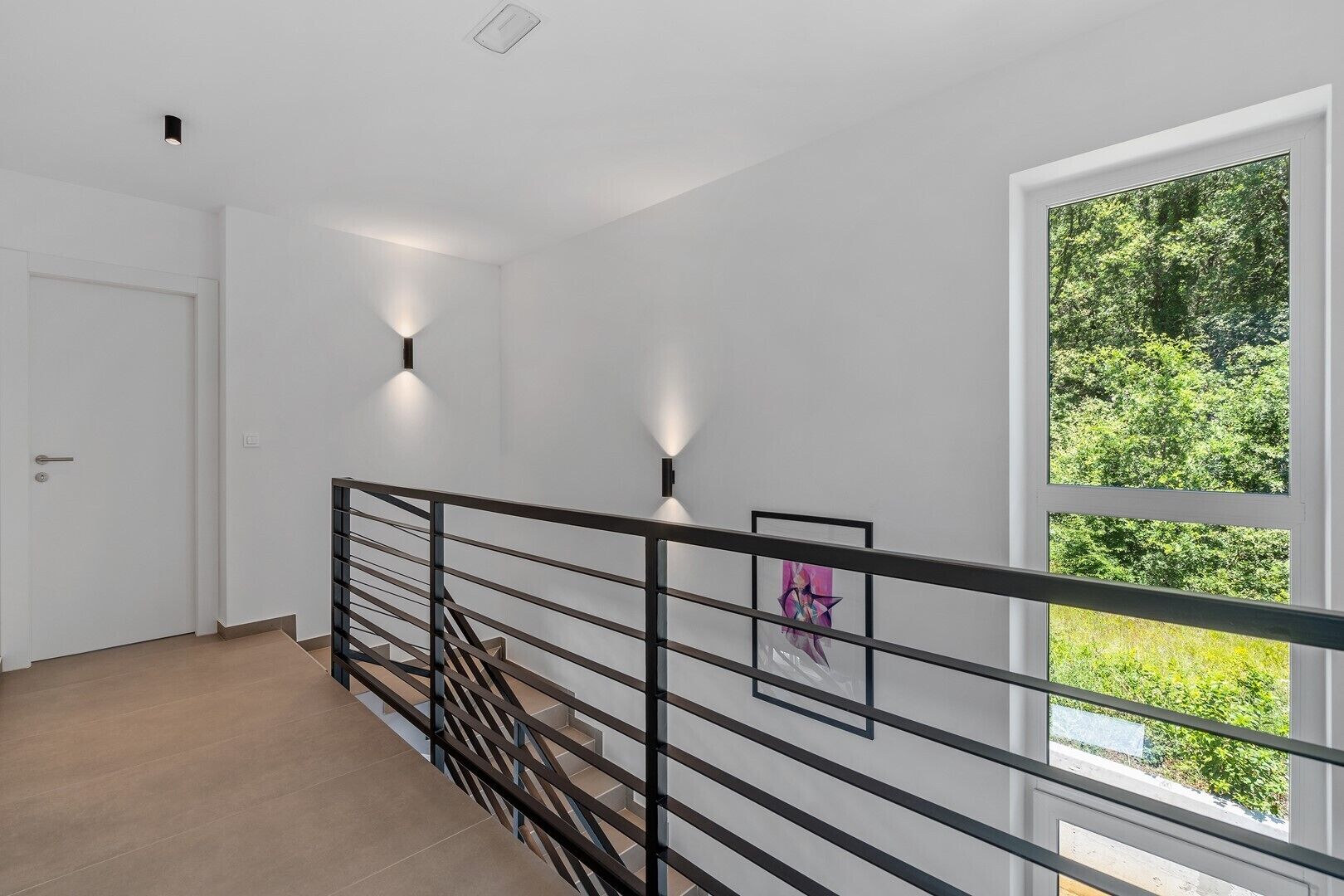 Un intérieur moderne avec un escalier, des murs blancs et une grande fenêtre donnant sur un paysage verdoyant.