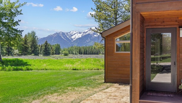 A wooden cabin with a glass door, set against a backdrop of mountains and a grassy field.