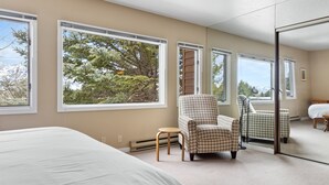 A bedroom with a large bed, two houndstooth patterned chairs, a small table, and a large window offering a view of trees and mountains.