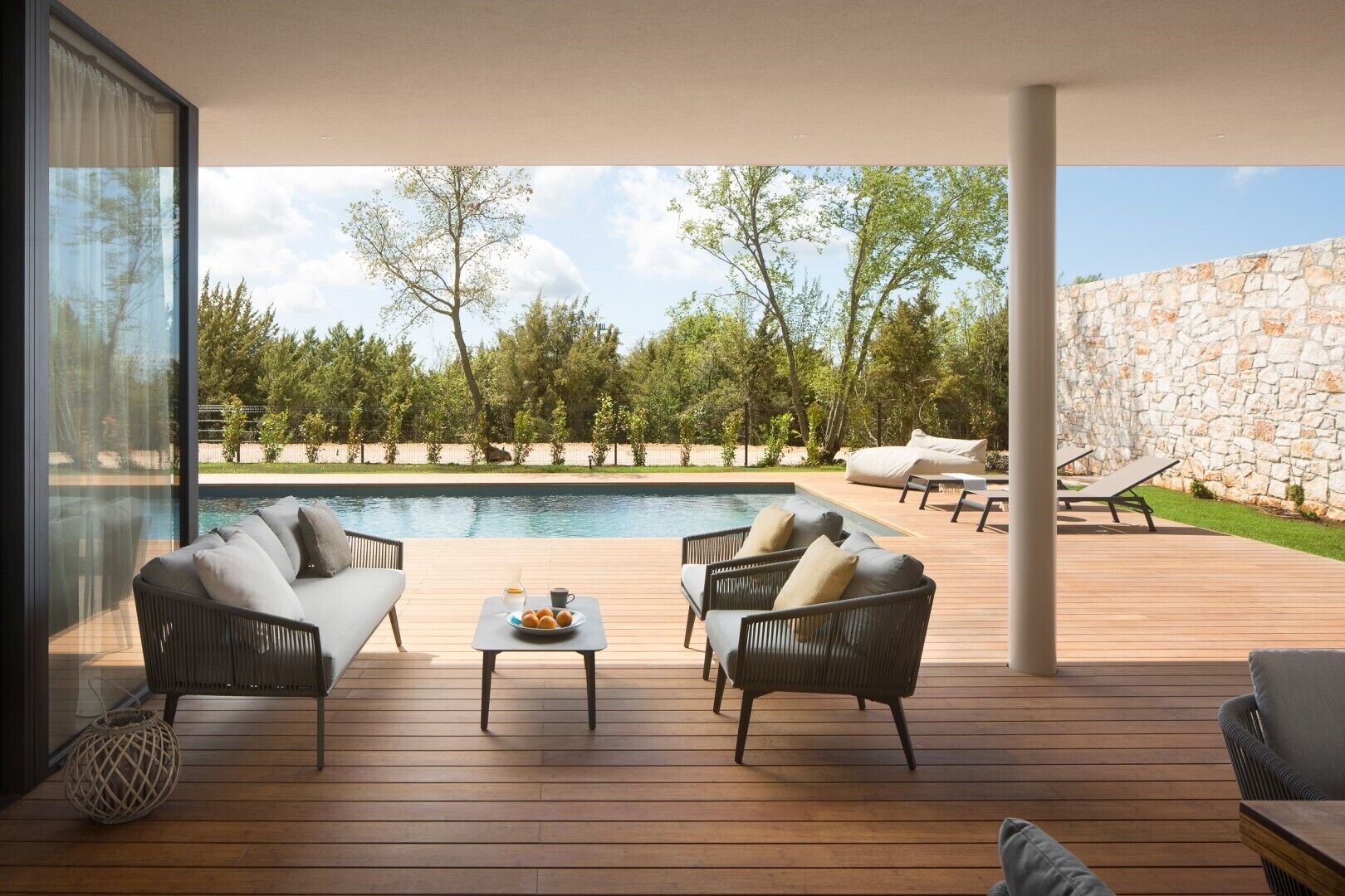 Une terrasse extérieure moderne avec une piscine, aménagée avec des fauteuils et une table en osier, et offrant une vue sur un mur de pierre et des arbres.
