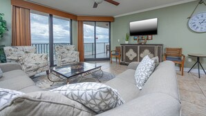 A spacious living room with a large sofa, a coffee table, and a television mounted on the wall. There is a balcony with a view of the ocean.