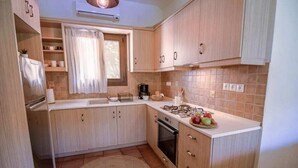 A compact kitchen with light wood cabinets, a built-in oven, and a window with curtains. - Luxury Villa Nefeli w Private Pool (Skiathos)