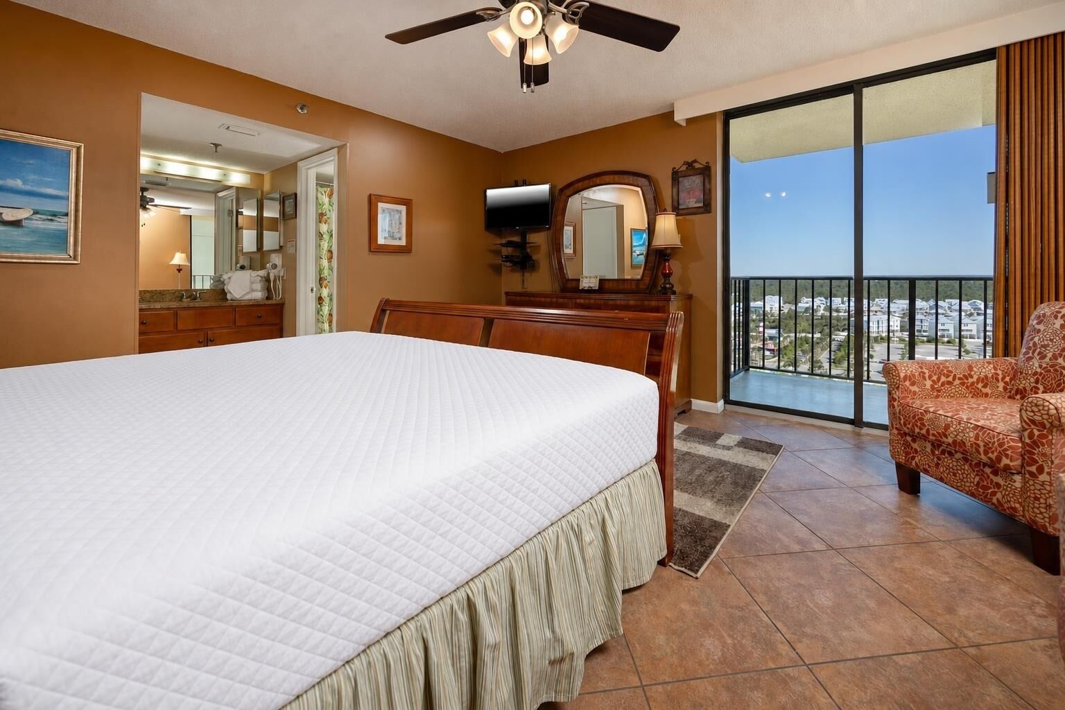 A hotel room with a bed, a chair, a TV, a balcony with a view, and a ceiling fan.