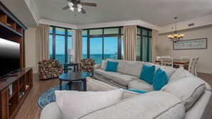 A living room with a large flat-screen TV, a sofa set, a coffee table, and a view of the ocean through large windows.
