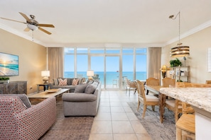 A spacious living area with a large window overlooking the sea, a ceiling fan, a dining area with a wooden table and chairs, and a modern kitchen in the background. - Phoenix Orange Beach Ii 703 2 Bedroom Condo (Orange Beach)